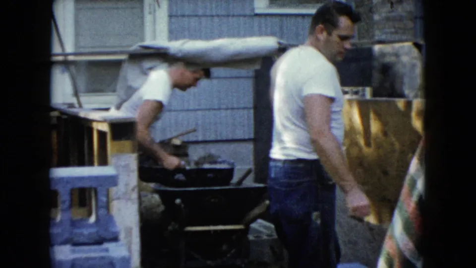 1960 Wauconda home-movie frame showing two adult men working near a wheelbarrow beside a residential exterior.