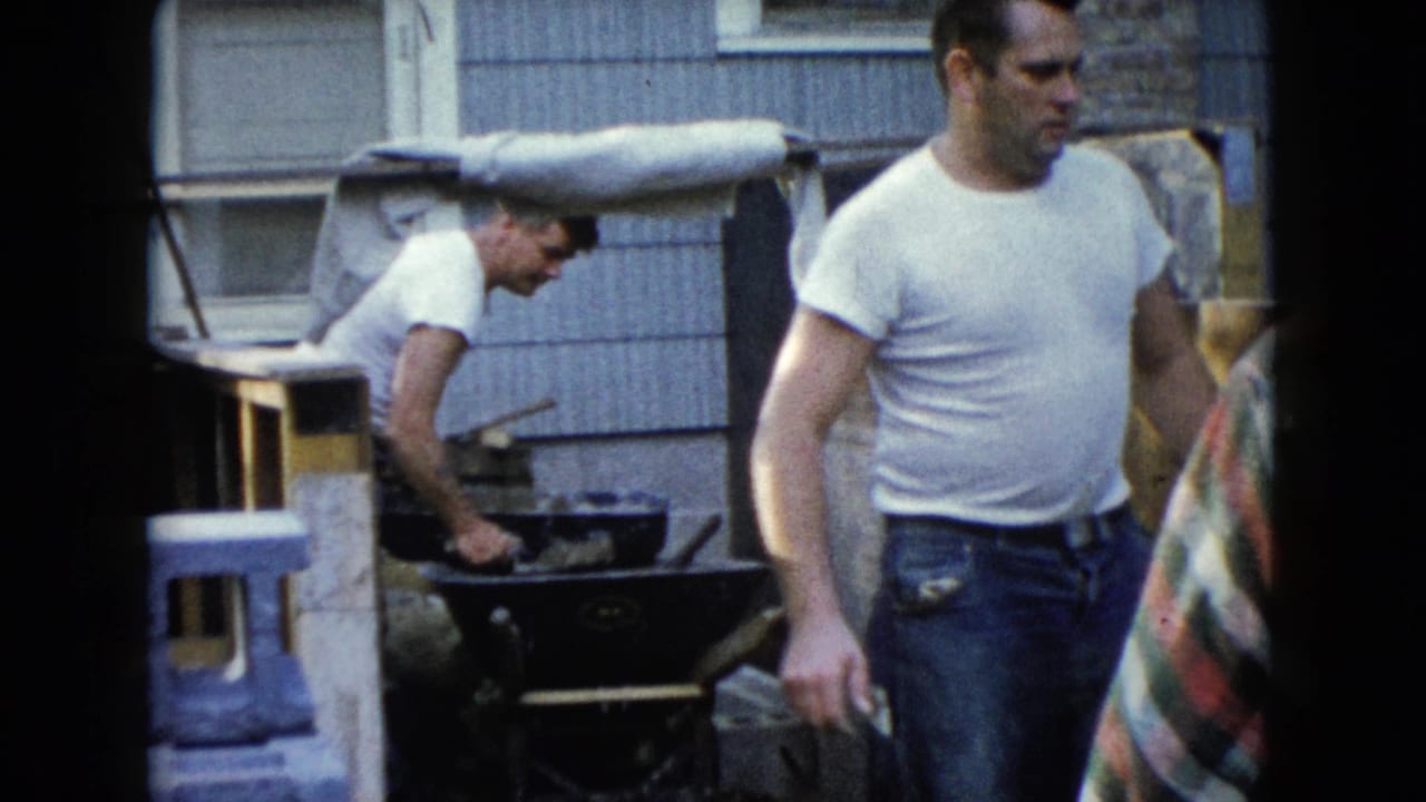 1960 Wauconda frame with one man passing close to camera and another bending over a wheelbarrow near house siding.