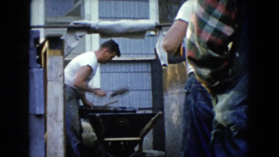 1960 Wauconda backyard frame showing a man working with a tool at a wheelbarrow, partly blocked by a nearby plaid-shirted figure.