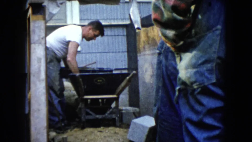 1960 Wauconda home-movie frame with a man leaning over a wheelbarrow beside yard materials and a residential exterior.