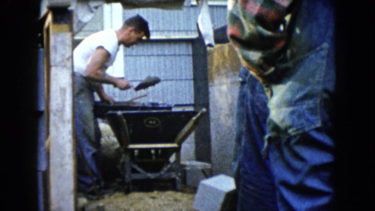 Young man mixing concrete in a wheelbarrow while another man stands nearby.