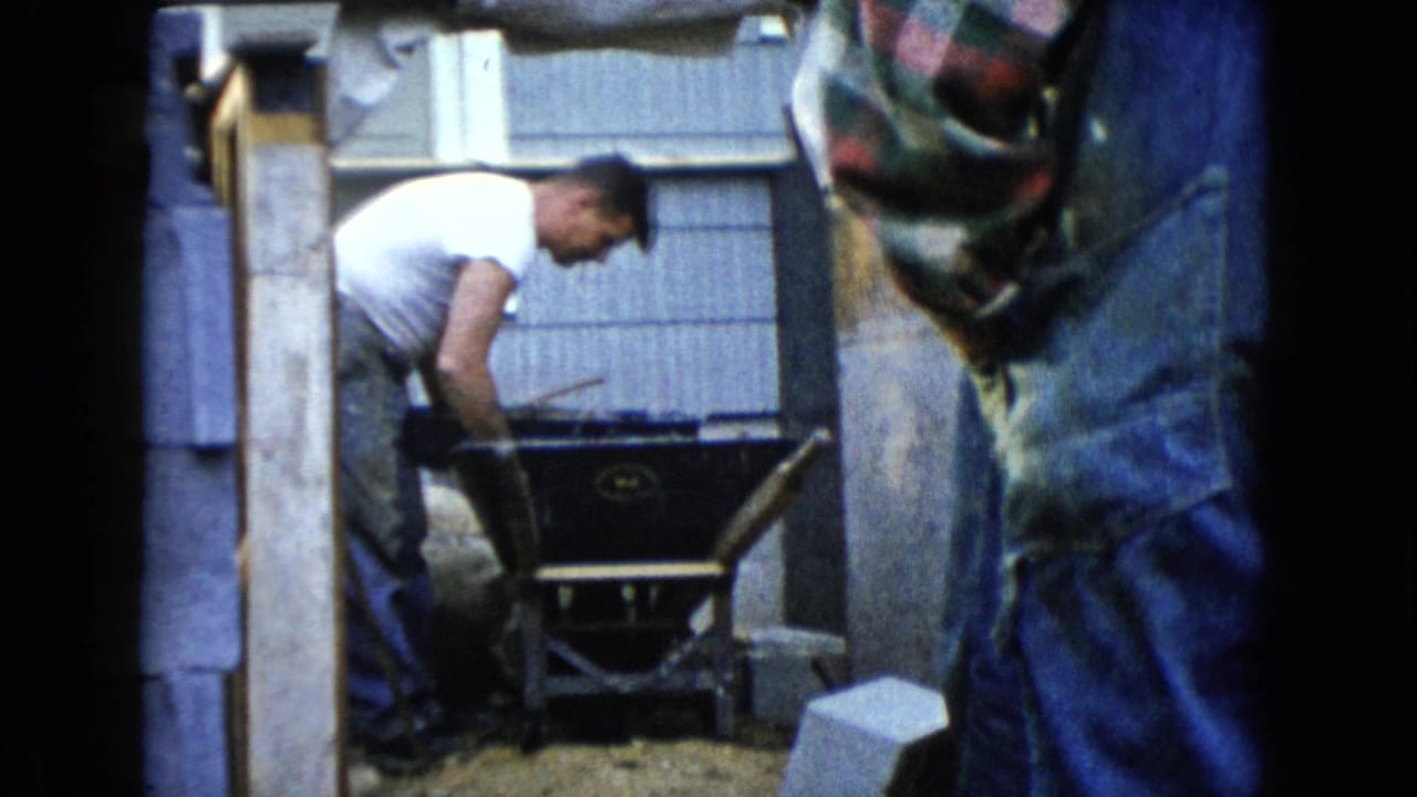 Man bends over a wheelbarrow with another adult partly blocking the foreground.
