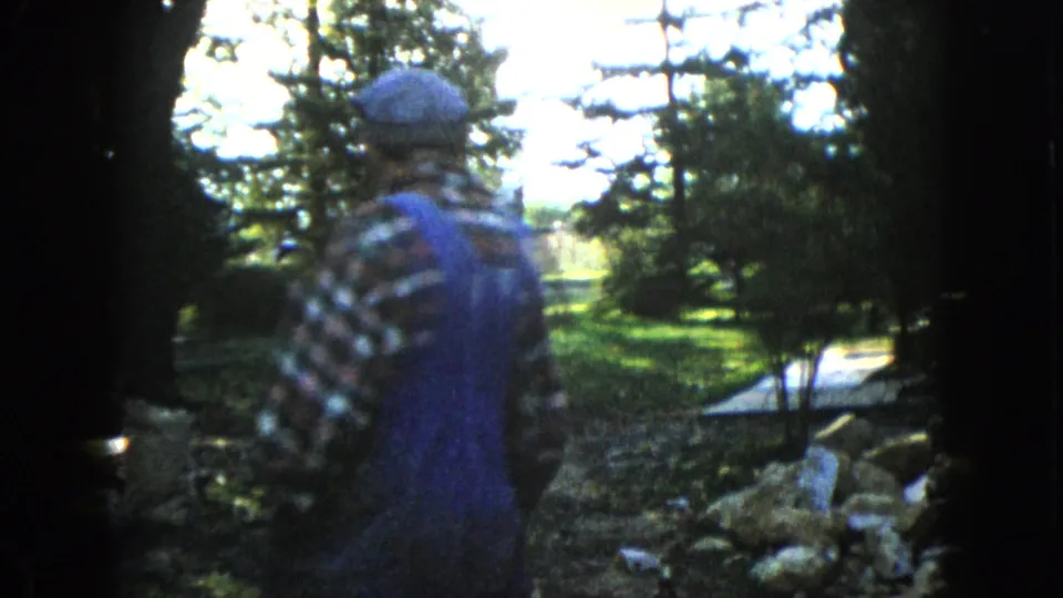 An older man in plaid and denim overalls walks away through a green Wauconda meadow or yard, seen in soft handheld 8mm footage.
