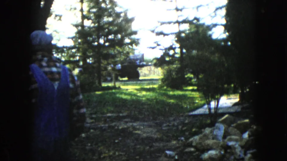 In Wauconda, Illinois around 1960, a person in blue overalls and plaid shirt walks away from the camera through a shaded garden toward a tractor.