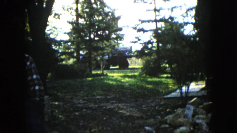 A partly visible figure moves at the edge of a shaded Wauconda garden while a tractor sits in the sunlit background among trees.