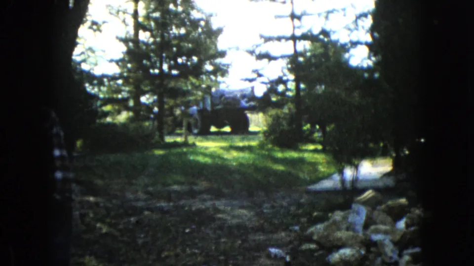 The camera looks through a shaded garden in Wauconda toward a tractor parked beyond trees, with the walking figure mostly out of frame.