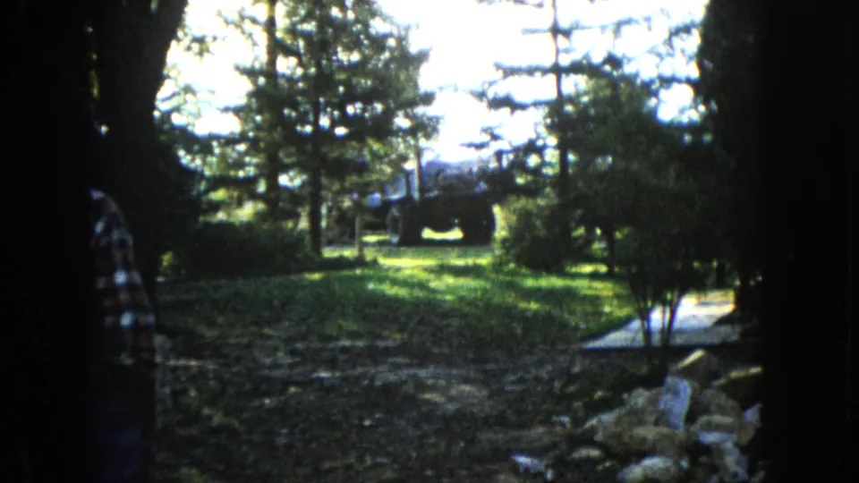 A person remains partly visible at the edge of a shaded Wauconda garden frame, with rocks, trees, and a tractor ahead in bright sunlight.