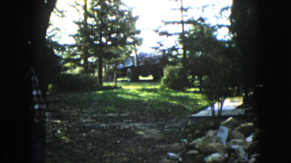 1960 8mm view in Wauconda, Illinois, looking through dark trees toward a sunlit grassy yard with rocks, shrubs, and a distant dark vehicle-like form.