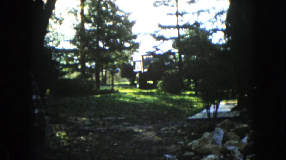 1960 Wauconda home-movie frame showing a shaded foreground, bright grassy yard, evergreens, shrubs, rocks, and a dark vehicle-like shape in the distance.