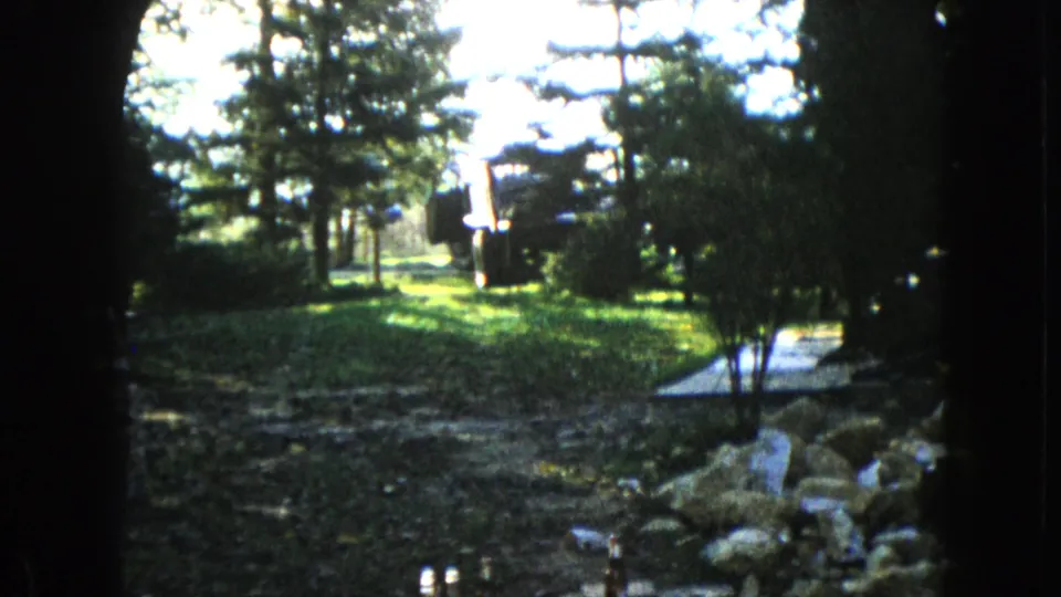 1960 8mm frame from Wauconda, Illinois, showing a high-contrast yard scene with grass, evergreens, shrubs, rocks, and bright sun patches.