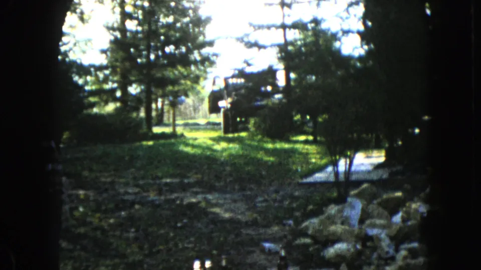 1960 Wauconda 8mm frame showing a shaded tree-framed yard, sunlit grass, shrubs, foreground rocks, a narrow path, and a distant dark shape.