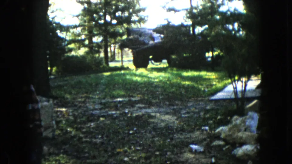 1960 Wauconda 8mm frame of a wooded yard, with dark foreground trunks framing grass, rocks, a paved path, shrubs, and a large dark object.