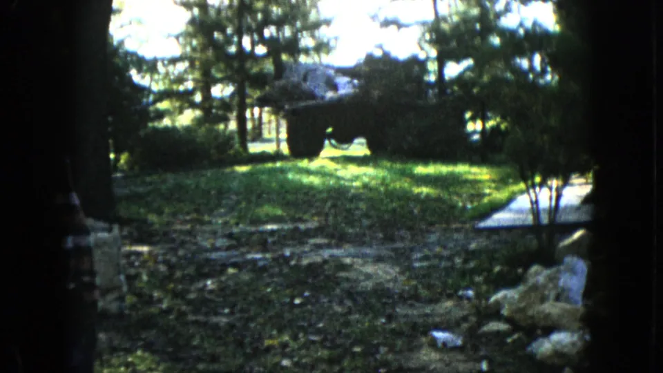 1960 Wauconda home-movie view across a shaded yard toward sunlit trees, rocks, shrubs, a path, and a large dark object in the background.