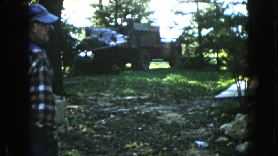 1960 Wauconda 8mm frame with an adult man in a plaid shirt and cap standing near dark tree trunks beside a wooded yard.