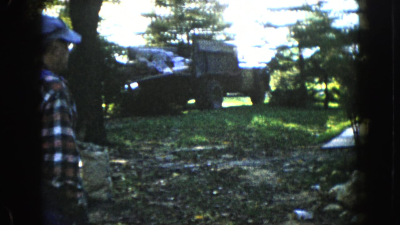In 1960 Wauconda, Illinois, a man in plaid watches a military-style truck loaded with supplies in a shaded wooded yard.