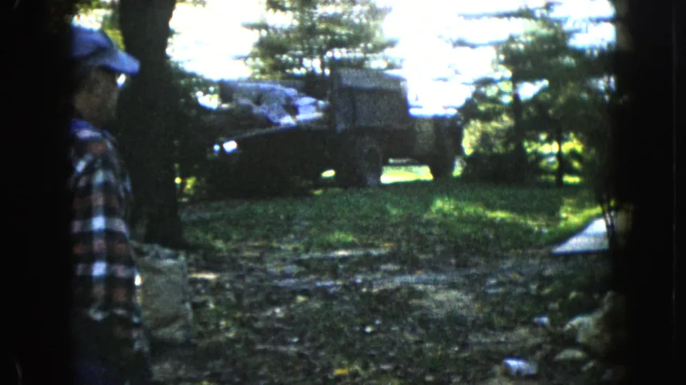 In 1960 Wauconda, Illinois, a man in plaid watches a military-style truck loaded with supplies in a shaded wooded yard.