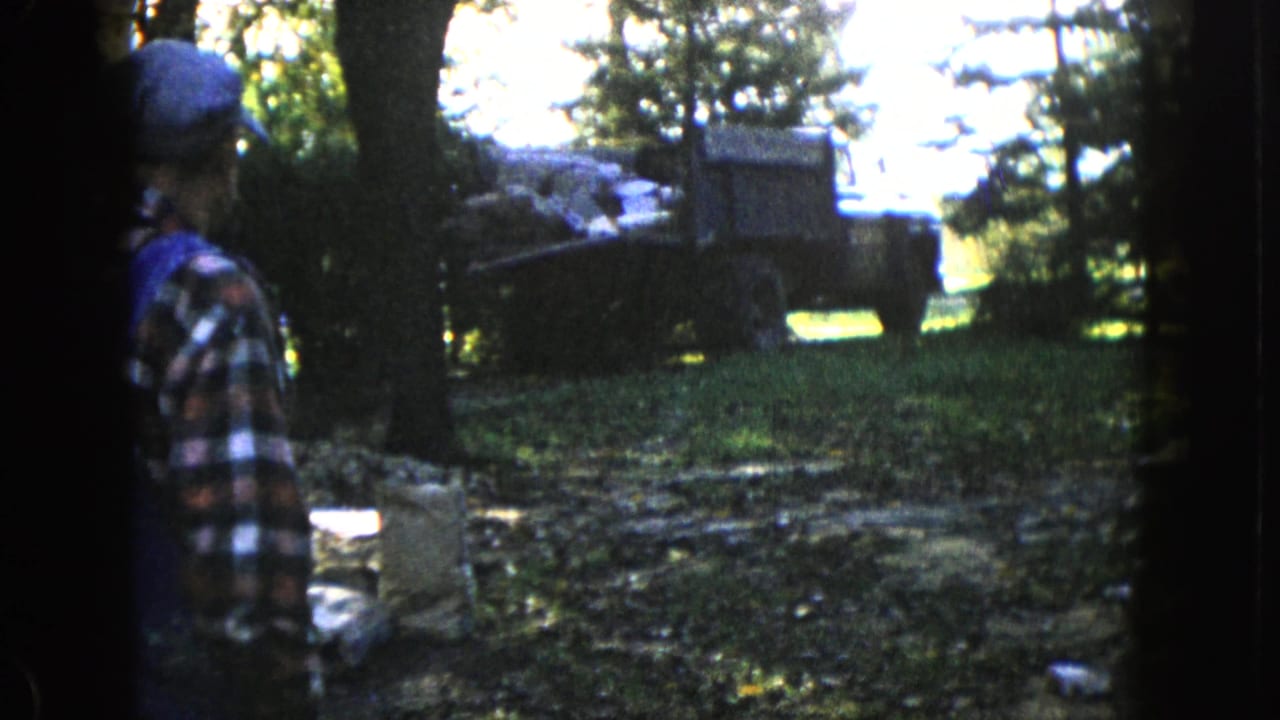 Man in plaid stands beside trees while a loaded truck is parked or backing in the background.
