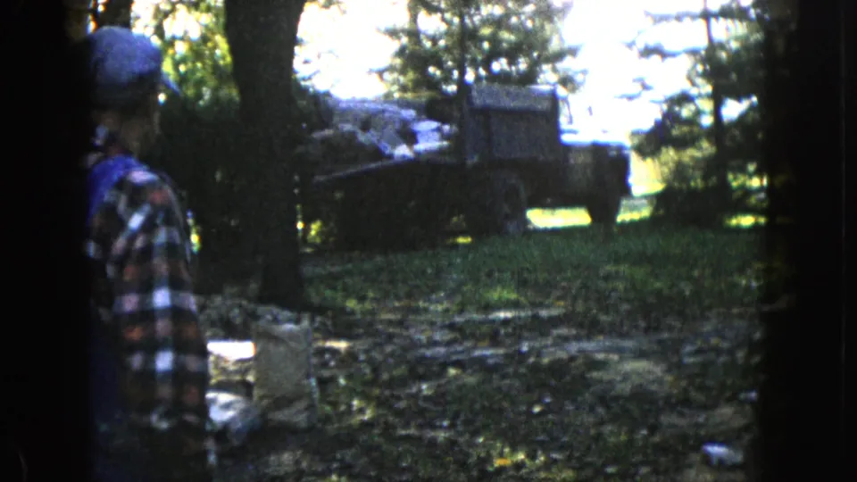 The 1960 Wauconda home-movie frame shows a man in plaid near trees with a supply-filled truck behind him.