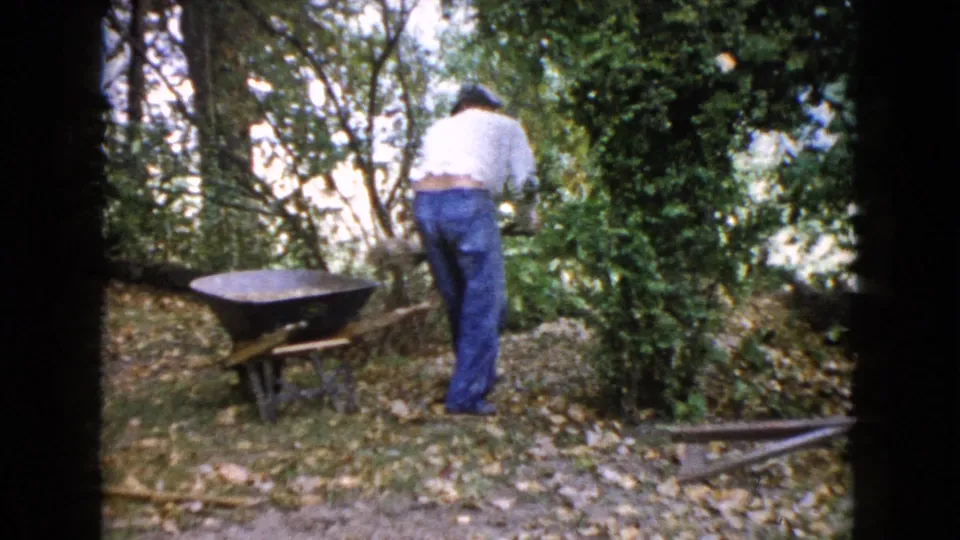 A man in a white shirt and blue jeans stands near shrubs and a wheelbarrow in the wooded Wauconda, Illinois yard.