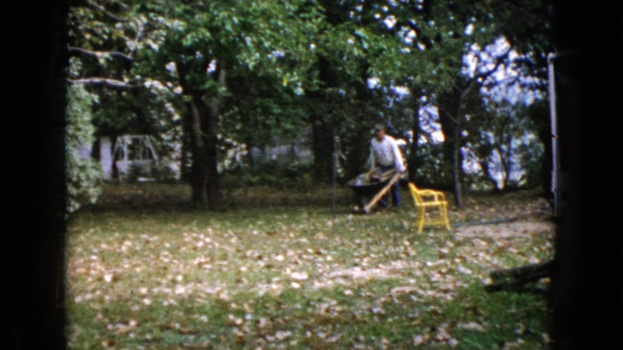 In Wauconda, Illinois home-movie footage from 1960, a man pushes a wheelbarrow across a leaf-covered backyard with trees and a yellow chair nearby.