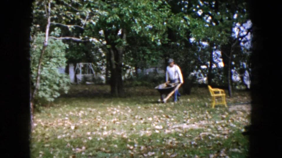 The 1960 Wauconda backyard scene shows the man guiding a loaded wheelbarrow across grass under trees, with a yellow lawn chair to one side.