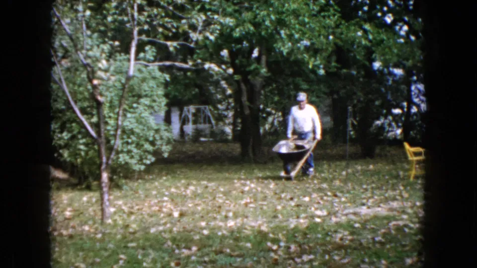 A man in a cap and light shirt pushes a wheelbarrow toward the camera in 1960 Wauconda home-movie footage, framed by trees and fallen leaves.