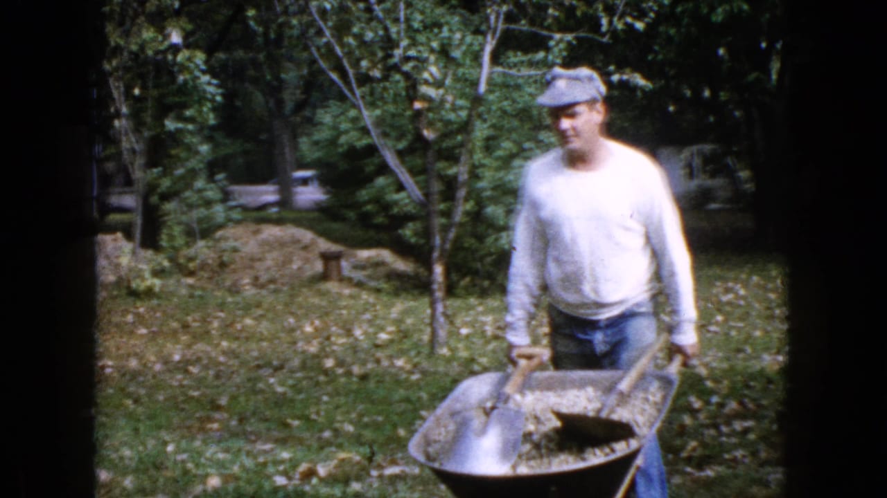 Man in a white shirt and cap pushes a wheelbarrow across a grassy yard.