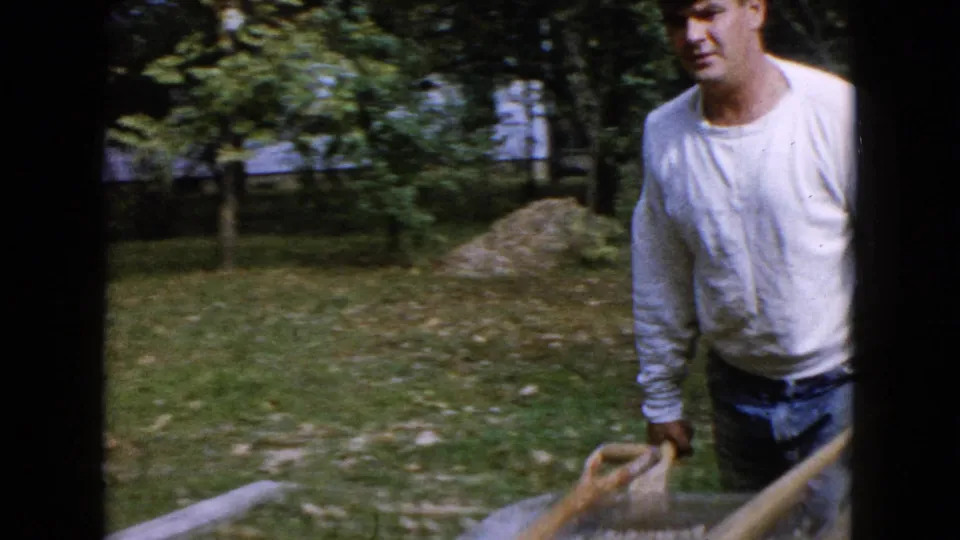 The man approaches with the wheelbarrow in a grassy residential yard, with trees and a dirt pile behind him.