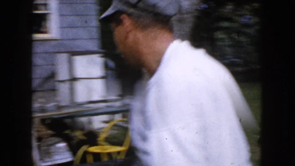 A close, slightly blurred side view shows the man in cap and white shirt passing the light-sided house in the 1960 home movie.
