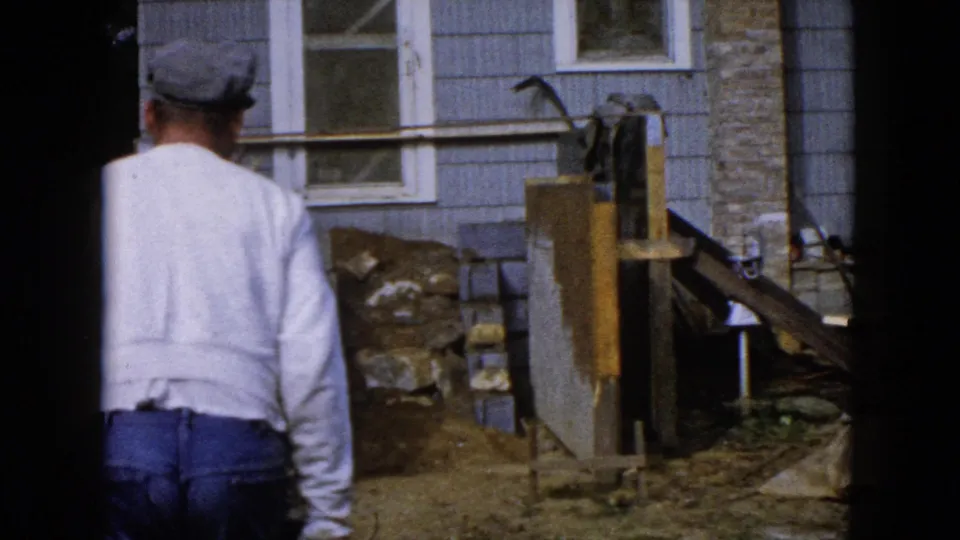 The man walks toward a light blue house exterior near a dirt pile, blocks, and construction equipment in Wauconda, Illinois.