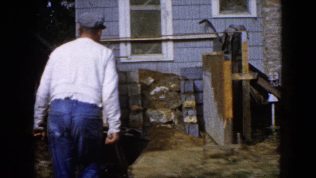 In 1960 Wauconda, Illinois footage, a man walks toward a residential construction area beside a blue-sided house.