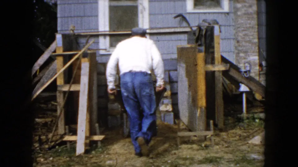 The man continues across the yard toward wood forms and stonework beside a blue-sided house in Wauconda, Illinois.