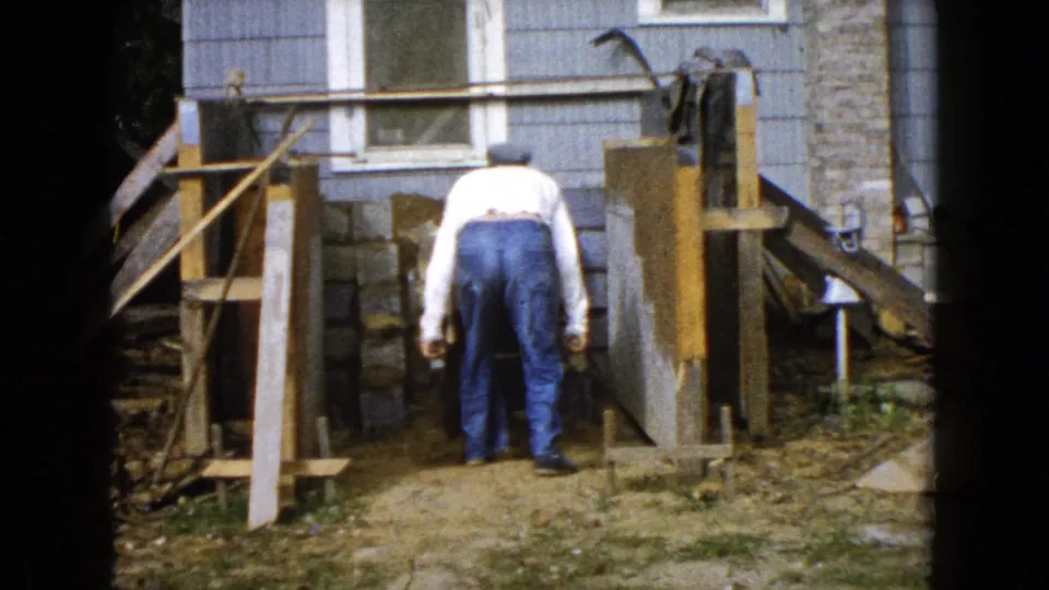 The man bends near the dirt work area, with wood supports, stonework, and house siding visible around him.