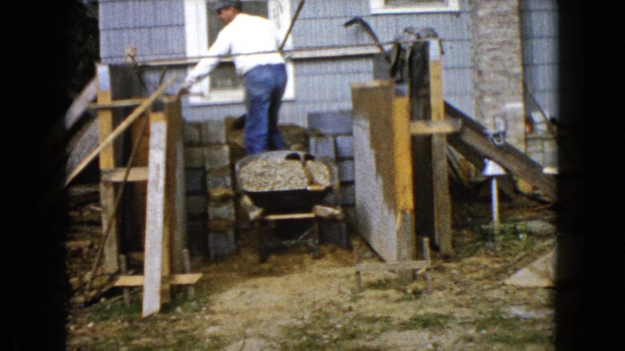 Man shoveling gravel near a wheelbarrow beside a house under construction.