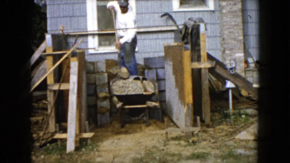 A man at the 1960 Wauconda construction project raises a shovel while standing near a wheelbarrow, wooden forms, and cinder blocks.