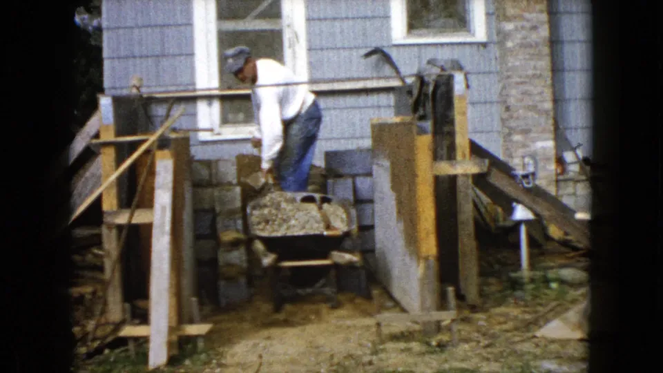 The man bends over the wheelbarrow during the 1960 Wauconda home construction work, with cinder blocks and wood framing visible behind him.