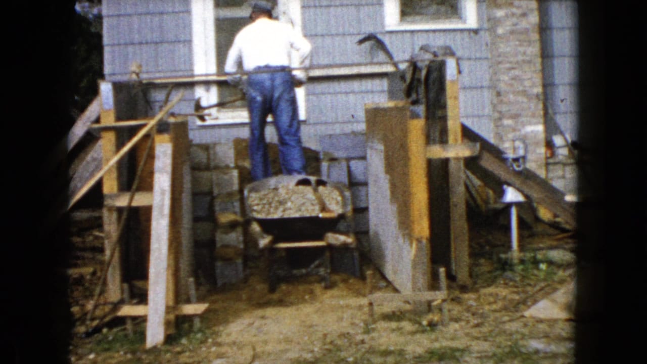 Man stands behind a gravel-filled wheelbarrow near a partly built porch structure.