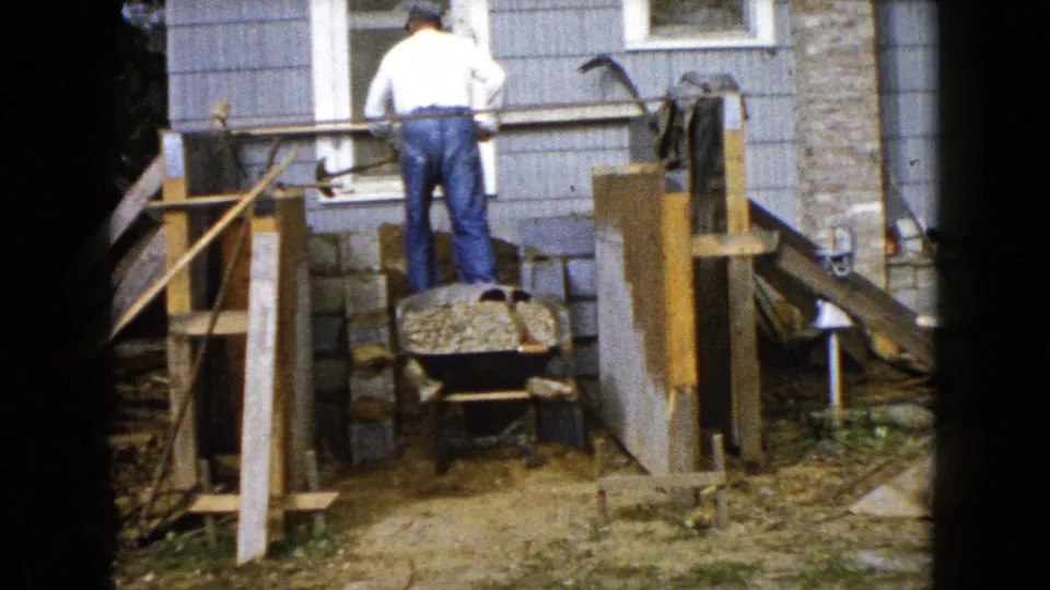 In Wauconda, Illinois circa 1960, a man in a white shirt and jeans works near a wheelbarrow, cinder blocks, and wooden forms at a house exterior.