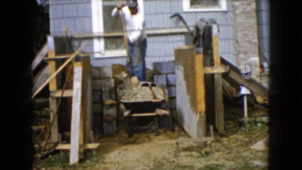 Man lifts a shovel near a wheelbarrow and cinder blocks beside a house.
