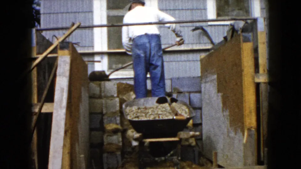 A rear view shows the man inside the narrow porch work area, with wooden forms, gravel, blocks, and the house wall visible around him.