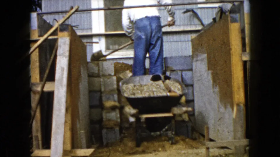 The closing frame shows the man from behind near the wheelbarrow, with cinder blocks, wood framing, dirt, and the house exterior still visible.