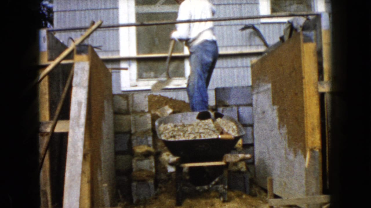 Man stands by a wheelbarrow of gravel near cinder blocks and wooden construction forms.