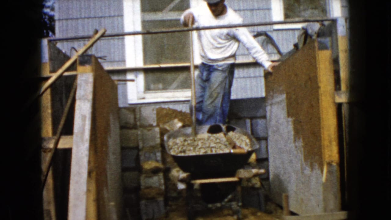 Man leans near a wheelbarrow and shovel between wooden forms at a house exterior.