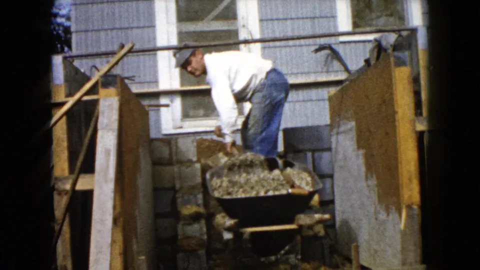 A man bends over a gravel-filled wheelbarrow near cinder blocks and wood forms in the Wauconda home construction scene.