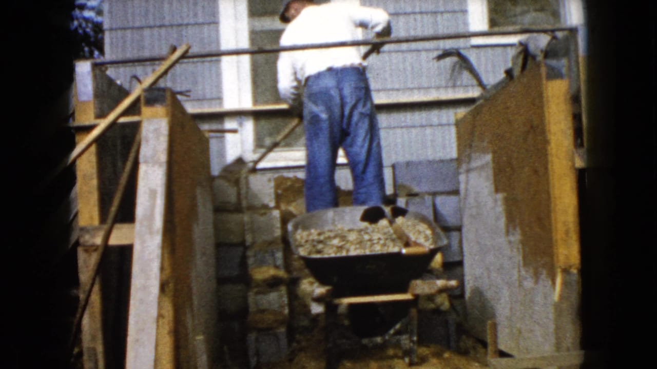 Man lays concrete blocks beside a house wall with a gravel-filled wheelbarrow nearby.