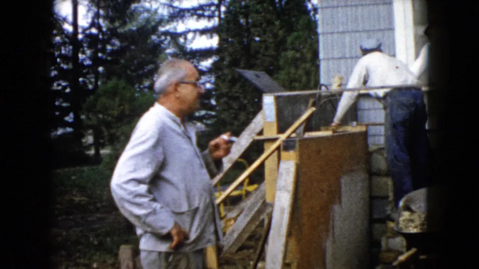 The home movie shows a man in profile holding a cigarette while another man works on concrete blocks beside a house exterior.