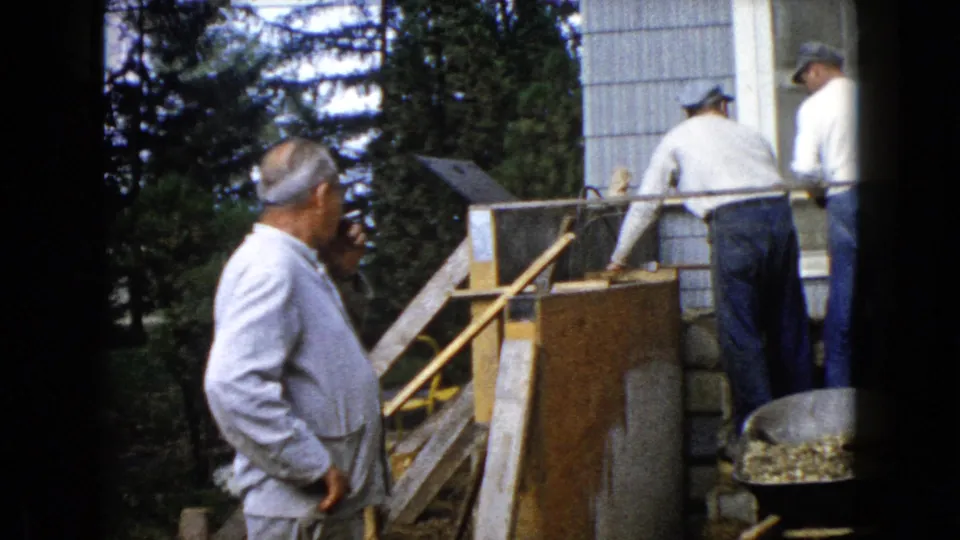 Two men appear beside a house foundation with concrete blocks, bracing, and a wheelbarrow in 1960 Wauconda, Illinois home-movie footage.