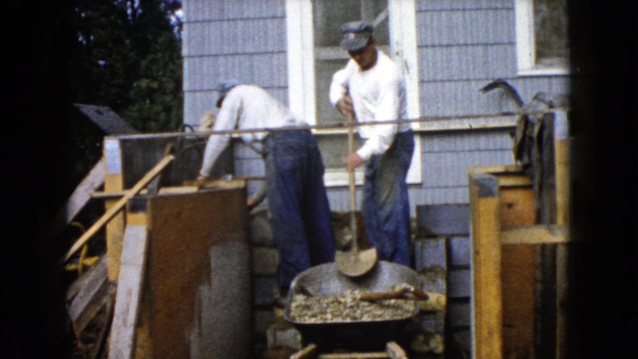 Two men stand and bend over masonry work beside a house with a wheelbarrow nearby.
