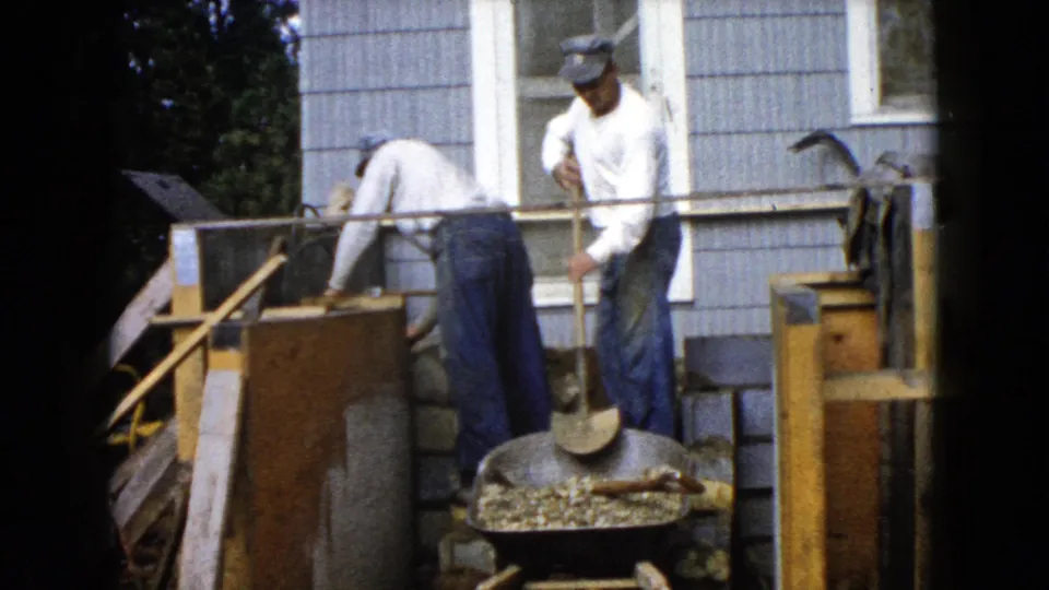 Two men in white shirts and work pants handle tools and materials beside a house during a 1960 Wauconda backyard construction scene.