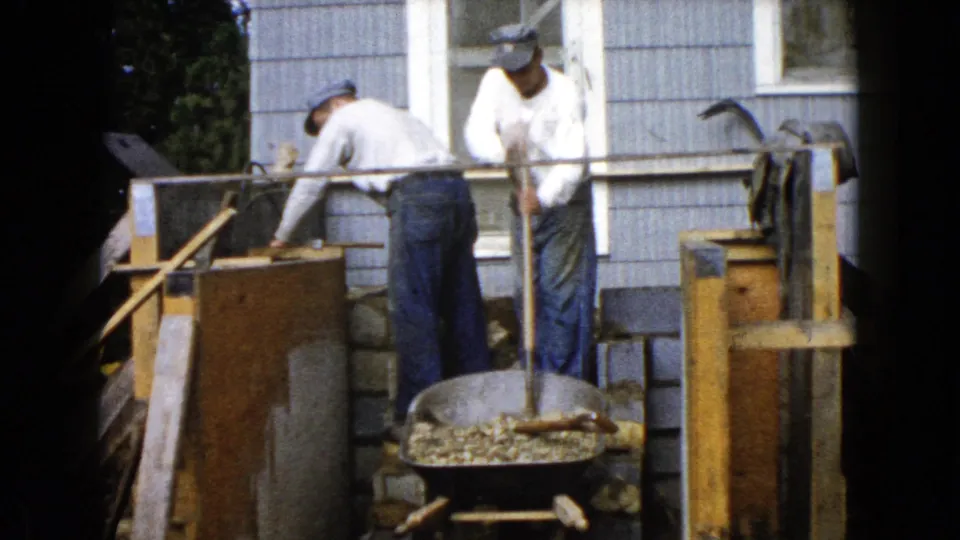 Men continue backyard masonry or construction work beside a Wauconda house, framed by an 8mm home-movie view.